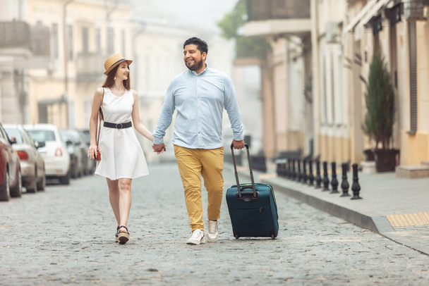 Κοινό, ρομαντικό ταξίδι. Αγαπώντας το διεθνές ζευγάρι των τουριστών με ταξιδιωτικές αποσκευές περπατούν κατά μήκος ενός ευρωπαϊκού δρόμου. Να περάσουμε χρόνο μαζί.. - Φωτογραφία, εικόνα
