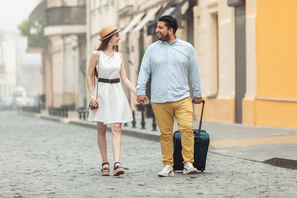 Joint, romantic trip. Loving international couple of tourists with travel luggage are walking along a European street. Spend time together. - Фото, изображение