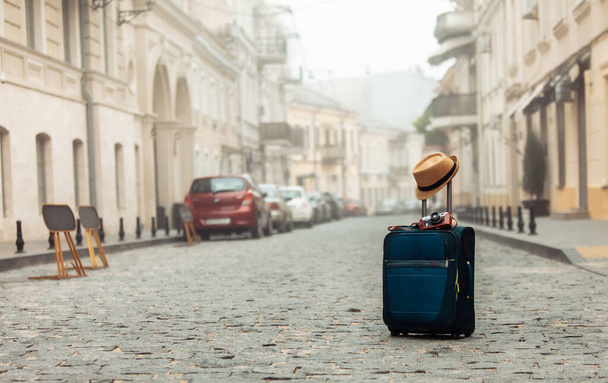 Ταξιδιωτικές αποσκευές με καπέλο και κάμερα στη μέση ενός άδειου ευρωπαϊκού δρόμου. Τουρισμός, Ταξιδιωτική έννοια - Φωτογραφία, εικόνα