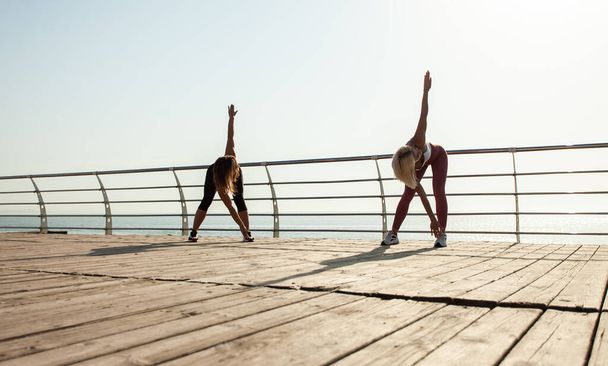 Two athletic women warming up doing exercises early morning on the beach - Photo, Image