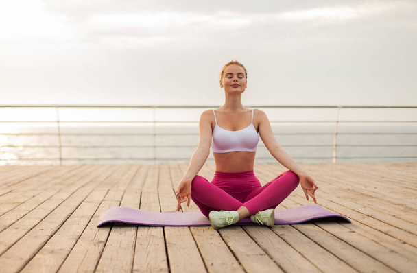Rothaarige Frau in Sportkleidung meditiert, während sie am Strand auf der Matte sitzt. Gesunder Lebensstil - Foto, Bild