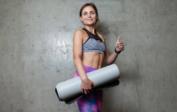 Jonge lachende fitte vrouw met yoga mat toont duim omhoog in de buurt van grijze muur. Gezonde levensstijl - Foto, afbeelding