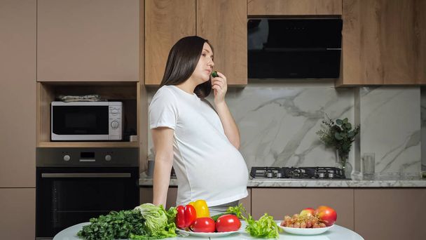 Woman eats vegetables stroking pregnant belly in kitchen - Foto, afbeelding