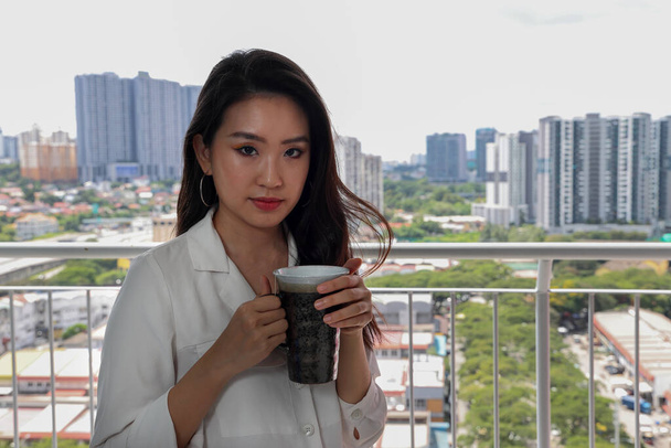 Beautiful young southeast Asian woman holding tea coffee cup near balcony window city scape background, looking relaxing thinking - Photo, Image