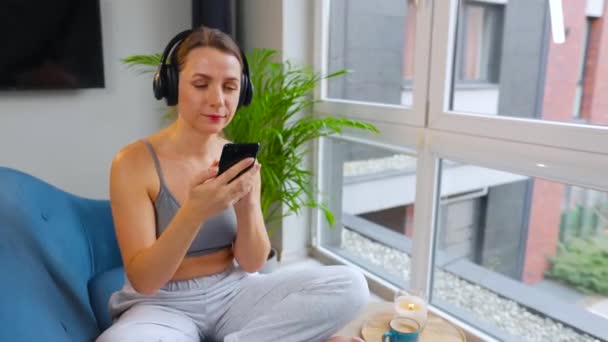 Γυναίκα με ασύρματα ακουστικά που ακούει μουσική και χρησιμοποιεί εφαρμογές για κινητά ή επικοινωνεί σε κοινωνικά δίκτυα σε smartphone που κάθεται σε καναπέ στο σπίτι - Πλάνα, βίντεο