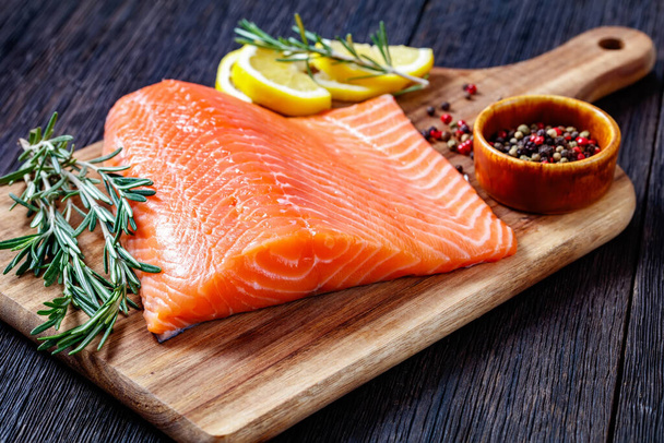 salmon fillet with peppercorn, rosemary and lemon on wooden cutting board on dark wood table, horizontal view from above, close-up - Foto, Imagem