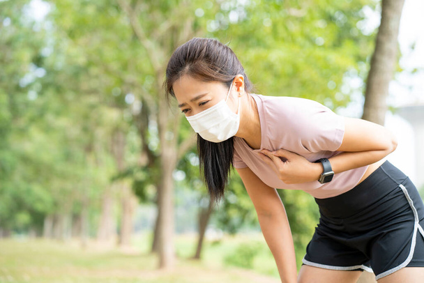 Asian woman runner wearing mask has chest pain and dizziness. Myocardial infarction may occur, see a doctor. Concept of the dangers of exercise. - 写真・画像