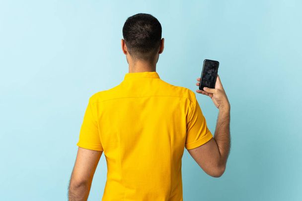 Jonge Colombiaanse man met behulp van mobiele telefoon geïsoleerd om blauwe achtergrond in achterpositie - Foto, afbeelding