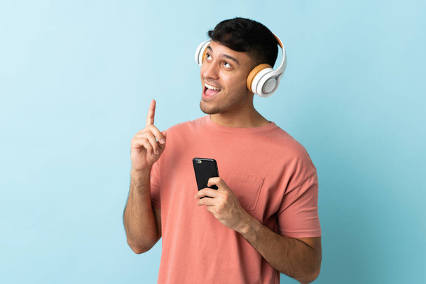 Молодий колумбійський чоловік, ізольований на синьому фоні, слухає музику мобільним телефоном і співає - Фото, зображення