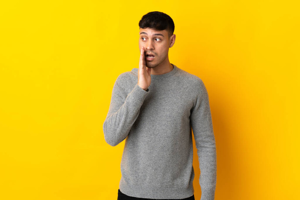 Young Colombian man isolated on yellow background whispering something with surprise gesture while looking to the side - Photo, Image
