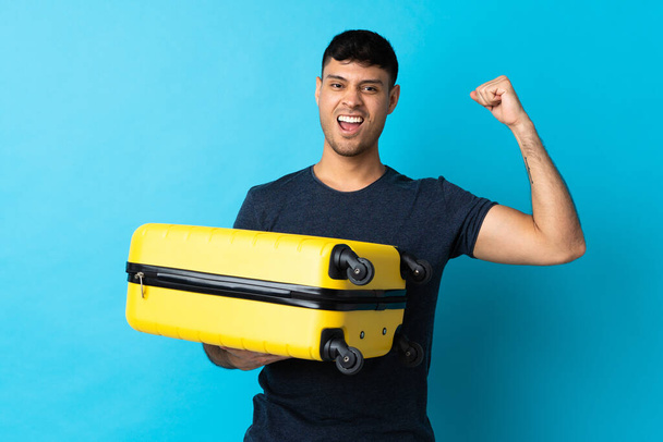 Young Colombian man isolated on blue background in vacation with travel suitcase - Photo, Image