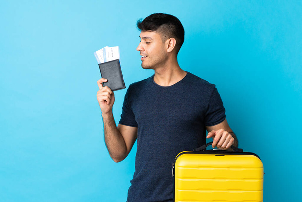 Genç Kolombiyalı bir adam mavi arka planda izole edilmiş, bavul ve pasaportla tatilde. - Fotoğraf, Görsel