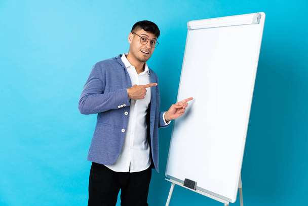 Young Colombian man isolated on blue background giving a presentation on white board and surprised while pointing side - Fotografie, Obrázek