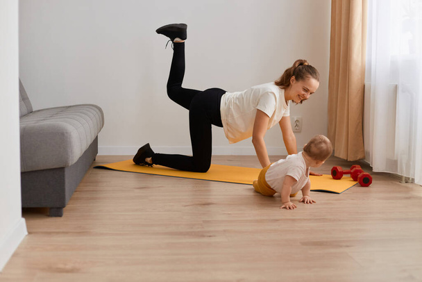 Side view portrait of fit woman doing bent knee donkey kick exercise in all fours position working out their buttocks at home with her little infant baby daughter, fitness and healthy lifestyle. - Фото, изображение