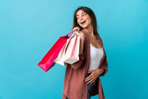 Young caucasian woman isolated on blue background holding shopping bags and smiling - Photo, Image