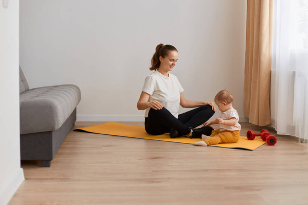 Full length portrait of happy smiling young mother does physical yoga exercises together with her baby daughter, looking at child, sitting in lotus pose, fitness at home, healthy lifestyle. - Photo, image