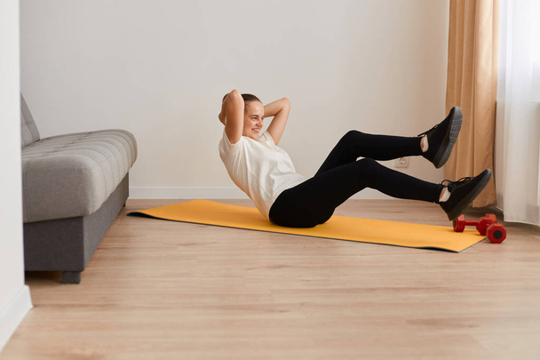 Fitness determined woman lying doing crunches at home, healthy female doing exercises on gym mat, happy young lady doing daily exercises lying on the floor in the living room. - 写真・画像