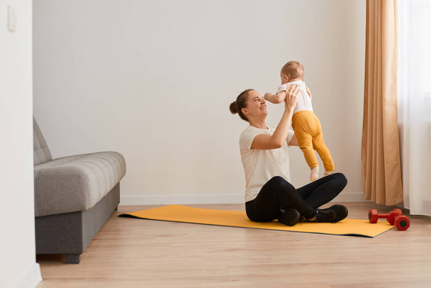 Portrait of athletic woman sitting in lotus pose on mat, wearing white t shirt and black leggins, posing in living room and throwing her baby daughter to the air while having rest, healthy lifestyle. - Photo, Image