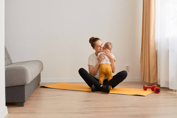 Full length portrait of sporty woman sitting in lotus pose on mat, wearing white t shirt and black leggins, training at home in living room and hugging her baby daughter while having rest. - Photo, Image