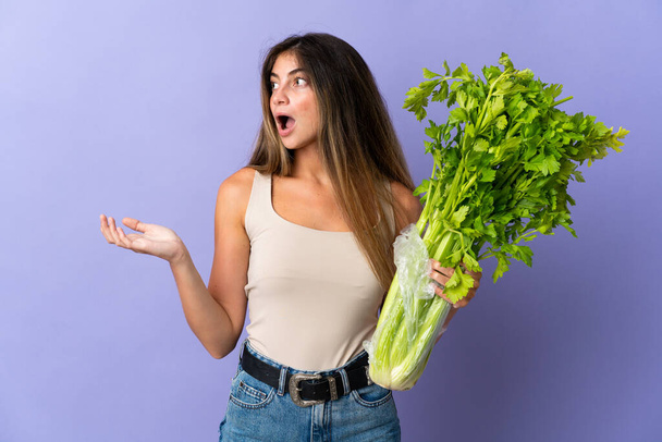 Young woman holding a celery isolated on purple background with surprise facial expression - Photo, Image