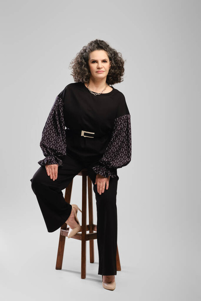 Senior woman with lush curly hair and black pantsuit sitting on tall chair in studioo over grey background - Foto, Bild