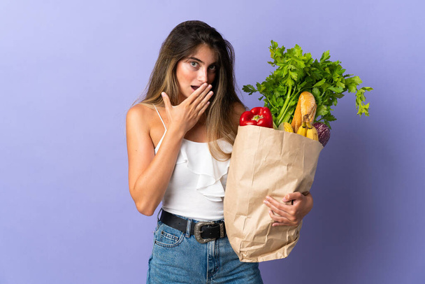 Young woman holding a grocery shopping bag happy and smiling covering mouth with hand - Photo, Image