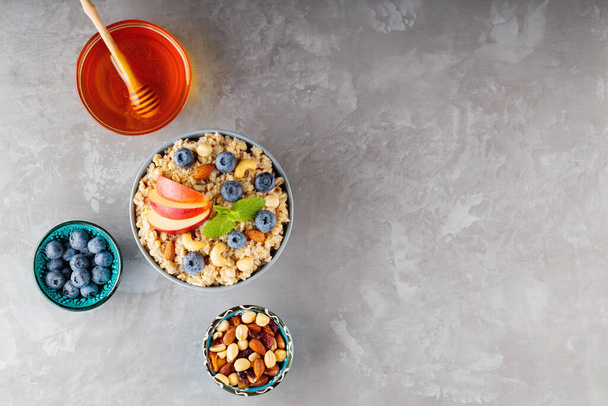 Healthy breakfast ingredients on a gray background. Oatmeal cereal, blueberries, nuts, apple slices and honey. Healthy breakfast concept. Top view. Copy space - 写真・画像