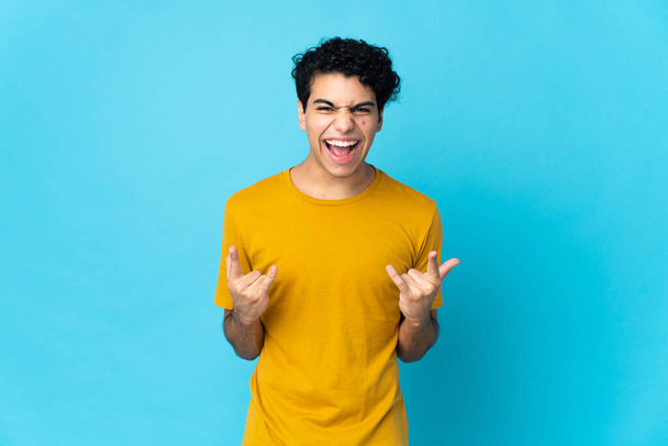 Young Venezuelan man isolated on blue background making horn gesture - Photo, Image