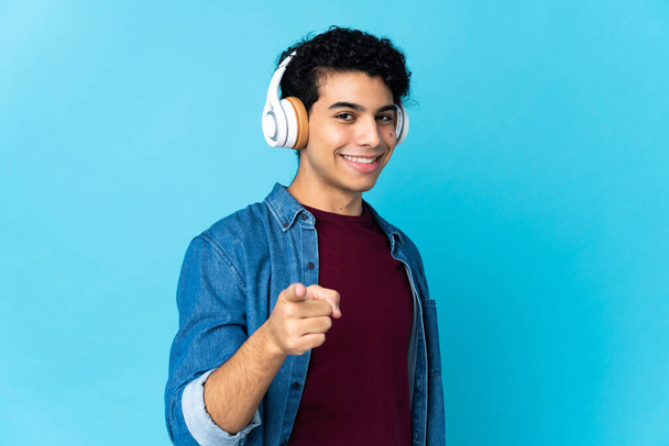Jeune homme vénézuélien isolé sur fond bleu écoutant de la musique et pointant vers l'avant - Photo, image