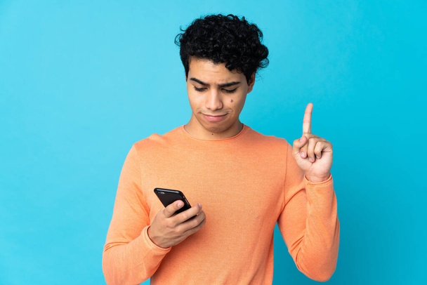 Венесуэльский мужчина изолирован на синем фоне с помощью мобильного телефона и пальца - Фото, изображение
