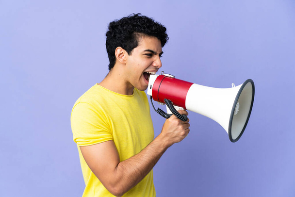 Young Venezuelan man isolated on purple background shouting through a megaphone to announce something in lateral position - Fotó, kép