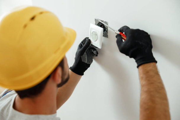 Professional electrician in uniform using screwdriver while installing electrical socket outlet after renovation work - Fotografie, Obrázek