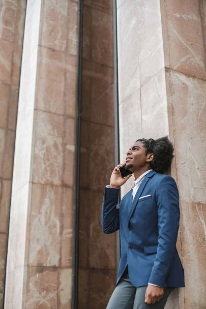 retrato vertical de empresário de etnia afro em uma chamada de trabalho, apoiando-se em uma parede, espaço de cópia vertical - Foto, Imagem