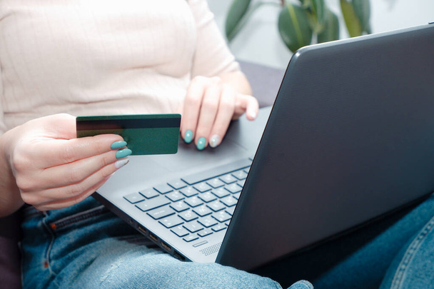 A woman at home sitting on a sofa holds a credit card in her hands and makes a payment using a laptop. happy online shopper. Woman using credit card to purchase merchandise on internet - Zdjęcie, obraz