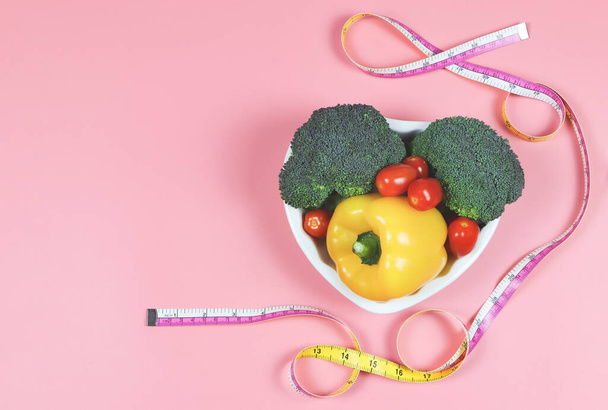 Bovenaanzicht of platte lay van groenten capsicum, broccoli en tomaat in hartvorm plaat met meetlint op roze achtergrond met kopieerruimte, gezonde levensstijl en gewichtsverlies concept. - Foto, afbeelding