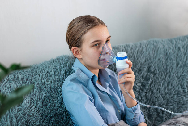 Teenage girl makes inhalation with a nebulizer equipment. Sick child holding inhalator in hand and breathes through an inhaler at home. Physical therapy for cold, flu and bronchial asthma - Fotografie, Obrázek