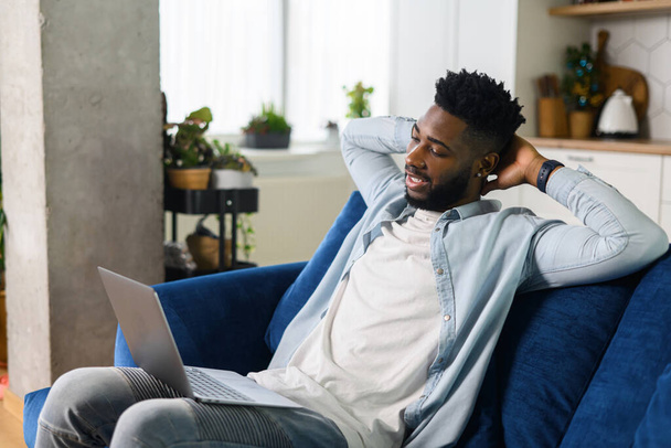 Cheerful multiracial man wearing casual wear sitting on the sofa and using laptop at modern home office, glad male entrepreneur - Zdjęcie, obraz