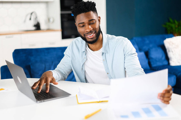 Executive multiracial male office employee doing paperwork while sitting at the desk with laptop in office. Focused and smiled man looking through sheets of documents - 写真・画像