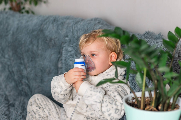 Cute baby boy makes inhalation with a nebulizer equipment. Sick child holding inhalator in hand and breathes through an inhaler at home. Physical therapy for cold, flu and bronchial asthma - Valokuva, kuva