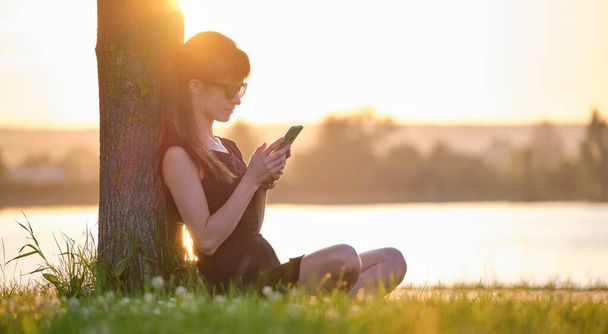 Młoda kobieta siedząca na zielonym trawniku nad brzegiem jeziora przeglądająca komórkę na świeżym powietrzu w ciepły letni wieczór. Koncepcja łączności i łączności ruchomej - Zdjęcie, obraz