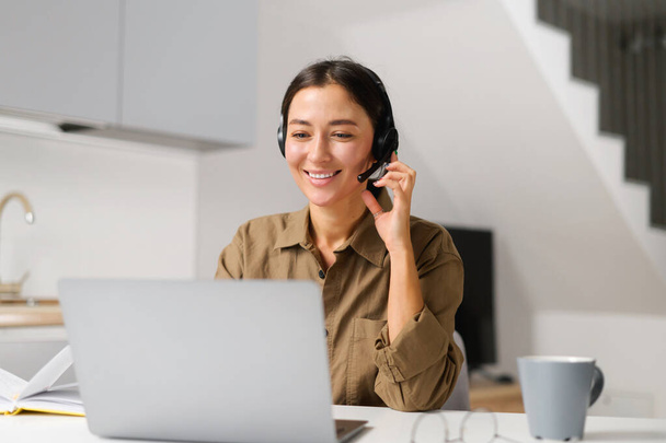 Young cheerful woman using headset for online talking sitting at the desk at home office. Pleasant female operator looking at the screen - Foto, Imagen
