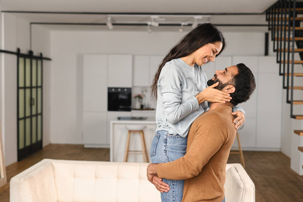 Cheerful Indian guy holding happy woman in arms while standing in apartment. Ethnic latin hispanic couple in love relocating in own flat. Move in new house - 写真・画像