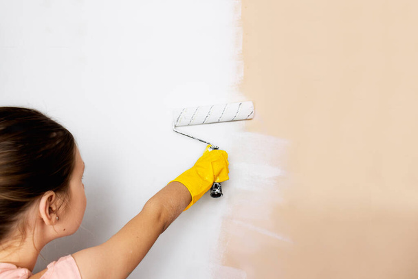 reparaciones en el hogar. pintar paredes con pintura. renovación. niña de guantes amarillos pinta una pared con un rodillo con pintura blanca. - Foto, imagen