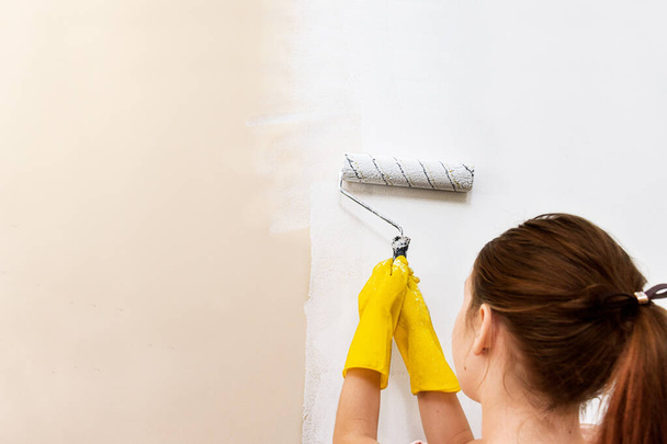 Hausreparaturen. Wände mit Farbe anstreichen. Renovierung. Mädchen in gelben Handschuhen bemalt mit einer Walze eine Wand mit weißer Farbe. - Foto, Bild