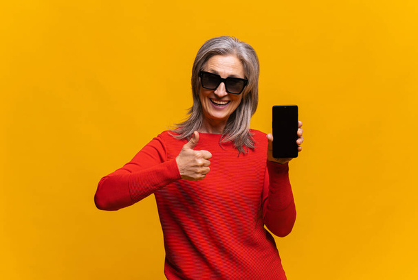 Horizontal shot of hilarious senior woman in red sweater holding smartphone with empty screen isolated. Middle-aged gray-haired lady showing thumb up, like sign - Fotoğraf, Görsel