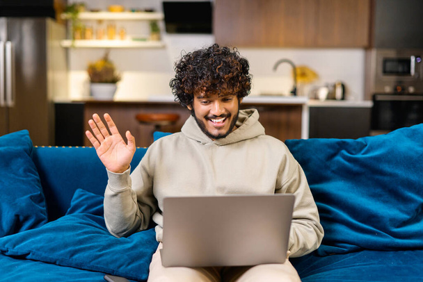 Video call. Cheerful attractive middle eastern guy is using laptop for video communication, while sitting at the sofa in modern flat and waving into webcam - Fotografie, Obrázek