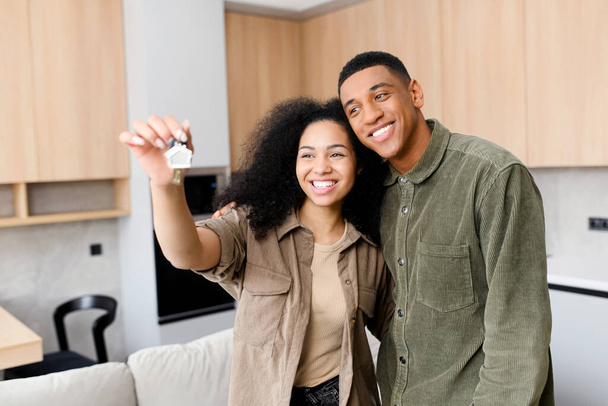 Happy young married couple holding a key ring in hand, hugging and looking at it with smile, while standing in the modern kitchen of their new home - Фото, изображение