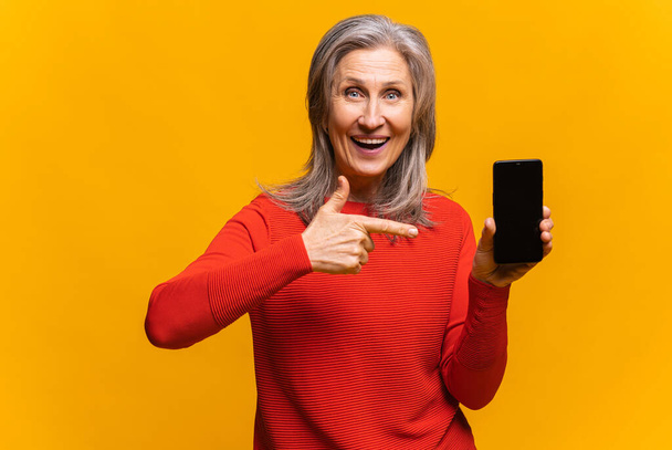 Positive woman looking at the camera and pointing to the smartphone with her finger isolated over yellow background. Advertises or recommends a new product, holiday sale concept - Zdjęcie, obraz