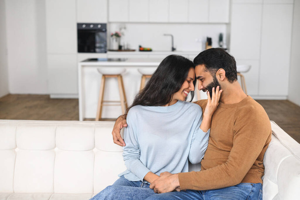 Happy Indian newlyweds moved in new apartment, sitting at the sofa in embraces, looking at each other. Multi ethnic couple in love rent a house together. Relocated in new home - 写真・画像