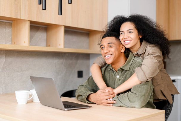 Cute multiracial couple enjoying leisure time at home with laptop. Happy woman and man in embrace looking at the screen and laughing, watching videos - Φωτογραφία, εικόνα